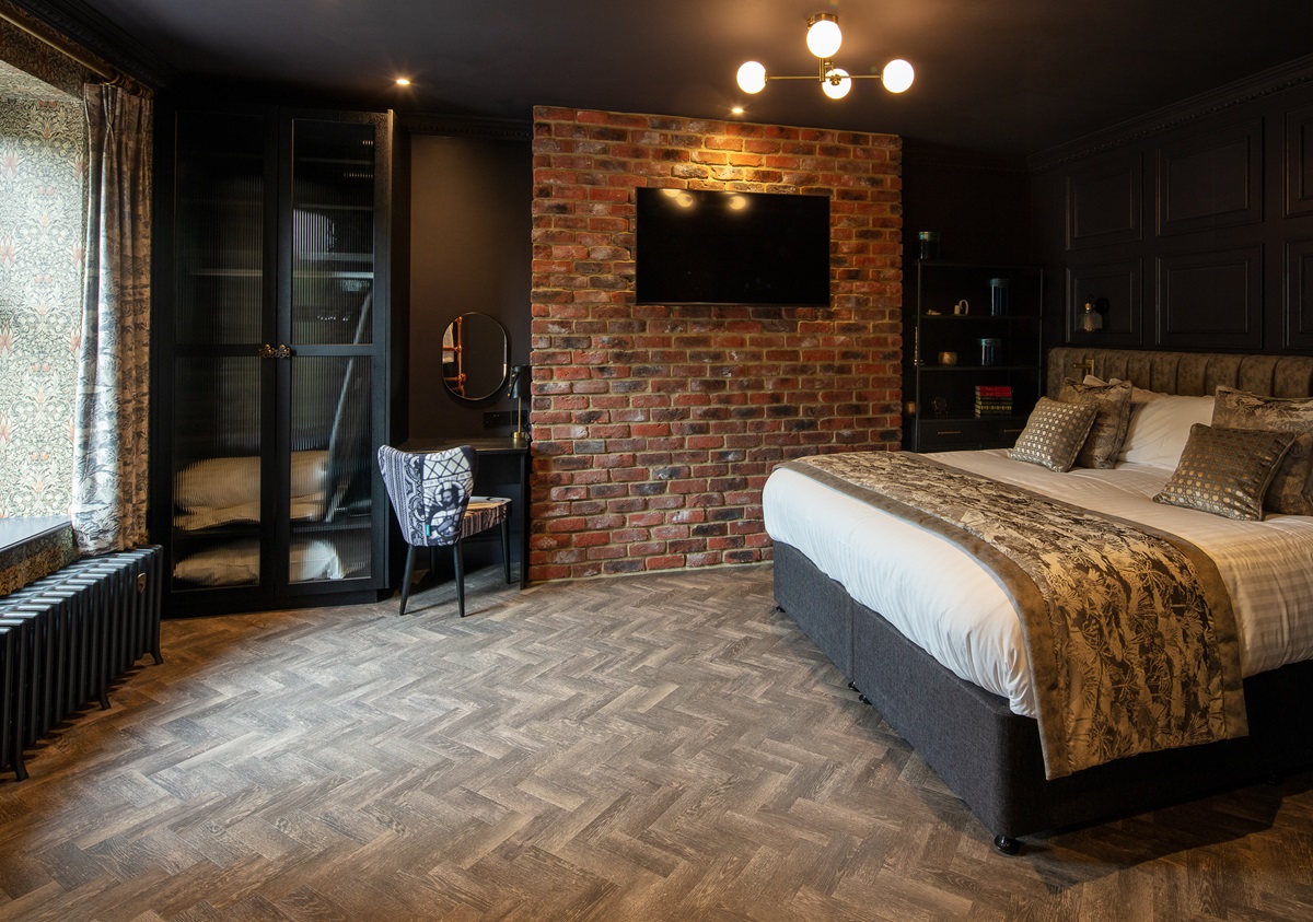 A dark guest room feels cosy and cocoon-like with dark walls, exposed brick, and herringbone flooring.