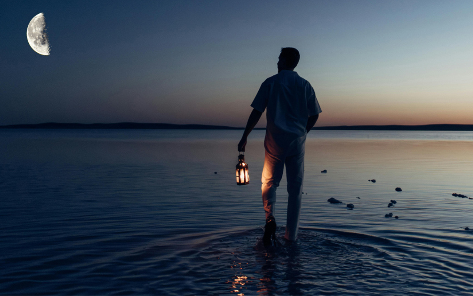 Man walking in the sea at dusk, beneath half moon