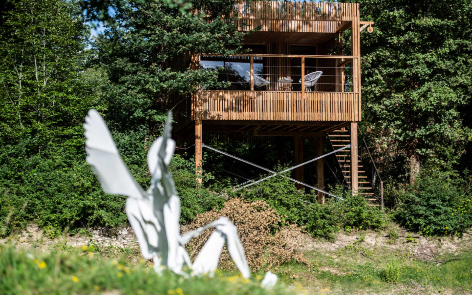 White metal angel sculpture, sitting on the ground with a shot of wooden Loire Valley Lodge in the background surrounded by treetops