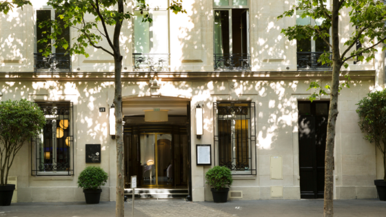 Pale facade of Le Narcisse Blanc Hotel, dappled with sunlight from trees lining the boulevard