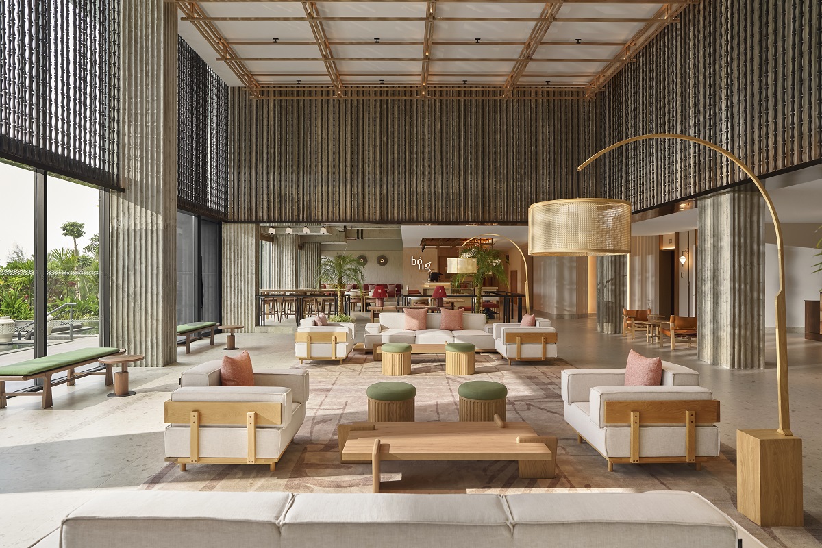 Spacious modern lobby of the NHAAN Resort & Spa Hoi An, Tapestry Collection by Hilton with beige sofas, green cushioned stools, wooden furniture, large floor lamp, and tall windows letting in natural light.