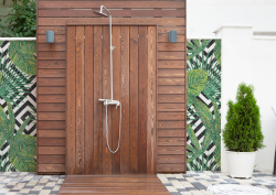 Outdoor shower area, with shower mounted on slatted wooden panel, surrounded by mosaic decorations which shows a green leaf pattern overlaid on a black and white geometric pattern