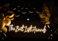 gold balloons and palm tree leaves surround a neon white and gold sign reading the brit list awards