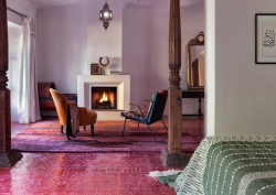 Guestroom at the Jnane Rumi in Marrakech, with pink tiled floor before wooden framed entrance to seating area with two armchairs infront of open fire. Floor turns to magenta and lilac woven rug