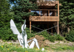 White metal angel sculpture, sitting on the ground with a shot of wooden Loire Valley Lodge in the background surrounded by treetops